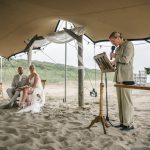 bruiloft op het strand met trouwambtenaar en bruidspaar dat zit en kijkt